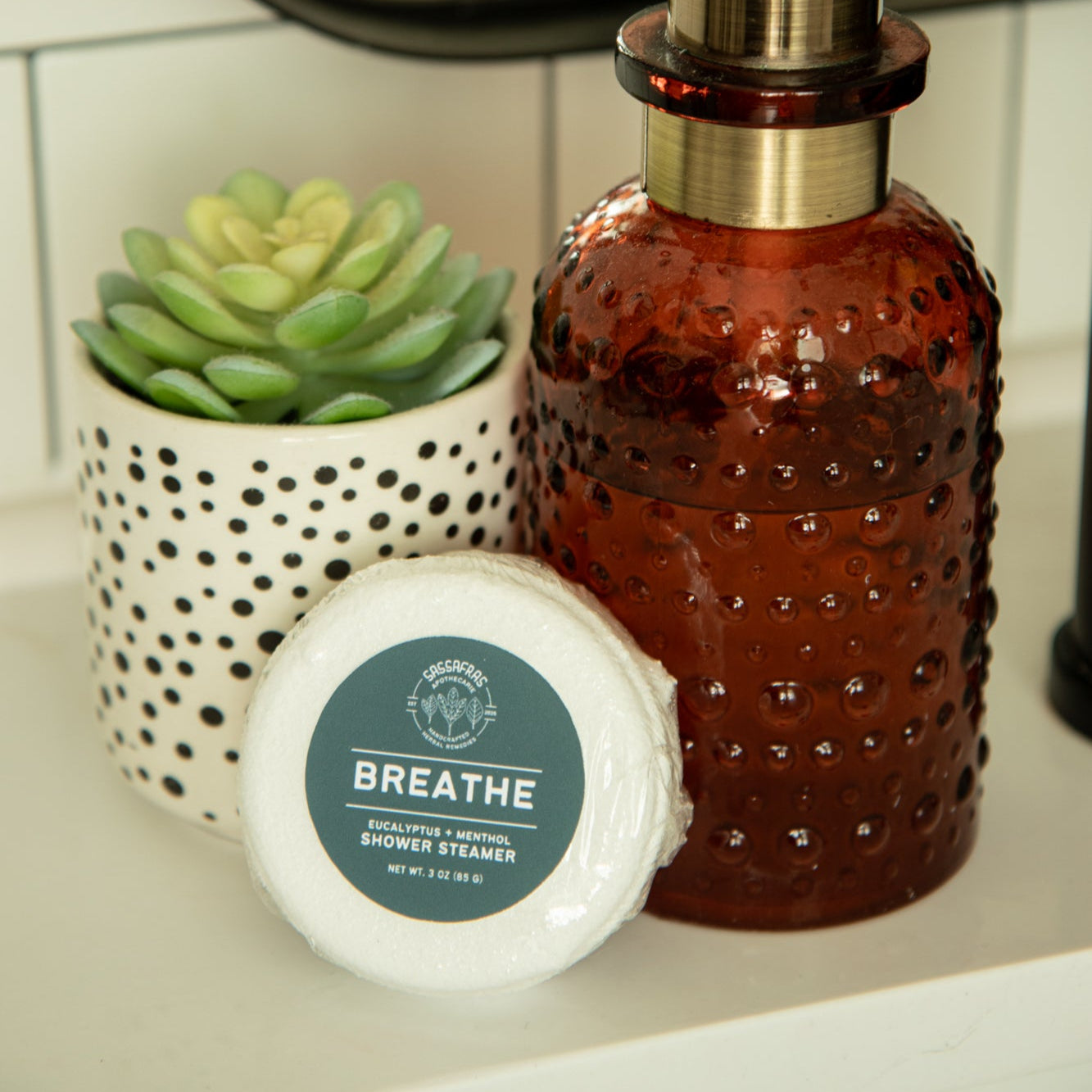 Brown glass soap dispenser with pump on a bathroom counter, next to a small plant and a shower steamer labeled 'Breathe'.